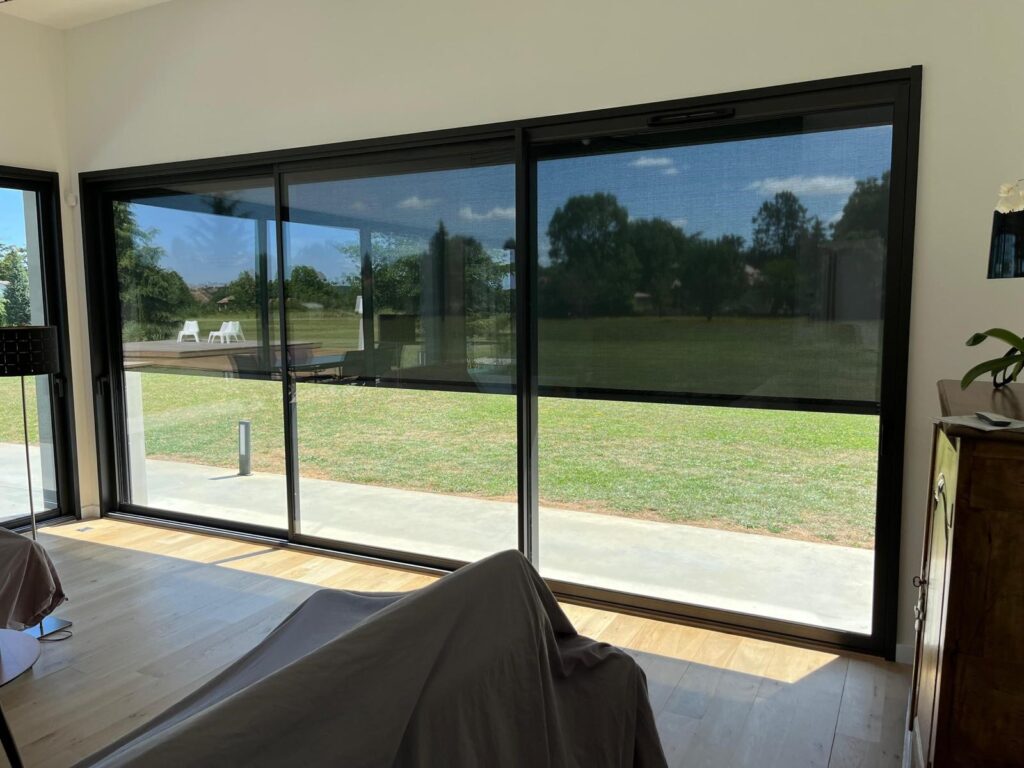 Porte-fenêtre au cadre noir pour une transition intérieure extérieure élégante. – Quercy Renov Porte-fenêtre coulissante à cadre noir, offrant une vue dégagée sur un patio extérieur moderne.
