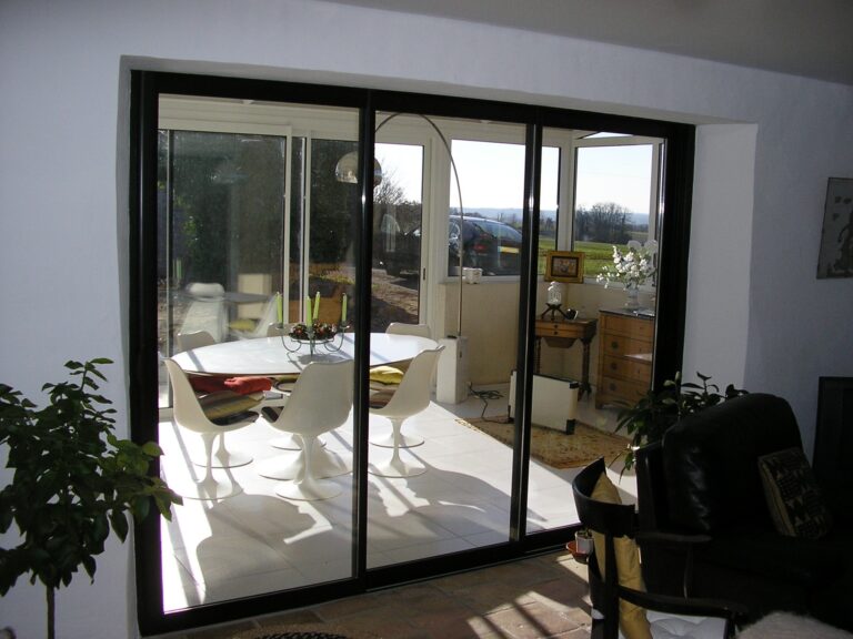 Porte-fenêtre moderne pour une lumière naturelle abondante. – Quercy Renov Grande porte-fenêtre en verre offrant une vue panoramique sur un patio lumineux et verdoyant.